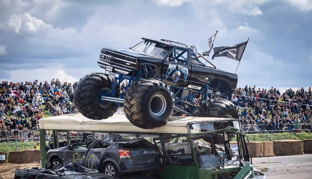 A monster truck on top of a bus and a row of cars