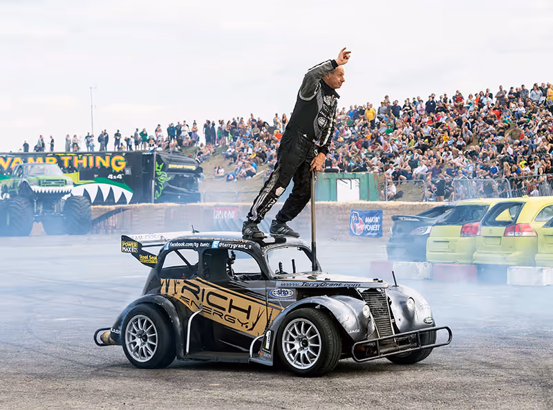 A stunt driver standing on the roof of a moving car in a cloud of tyre smoke